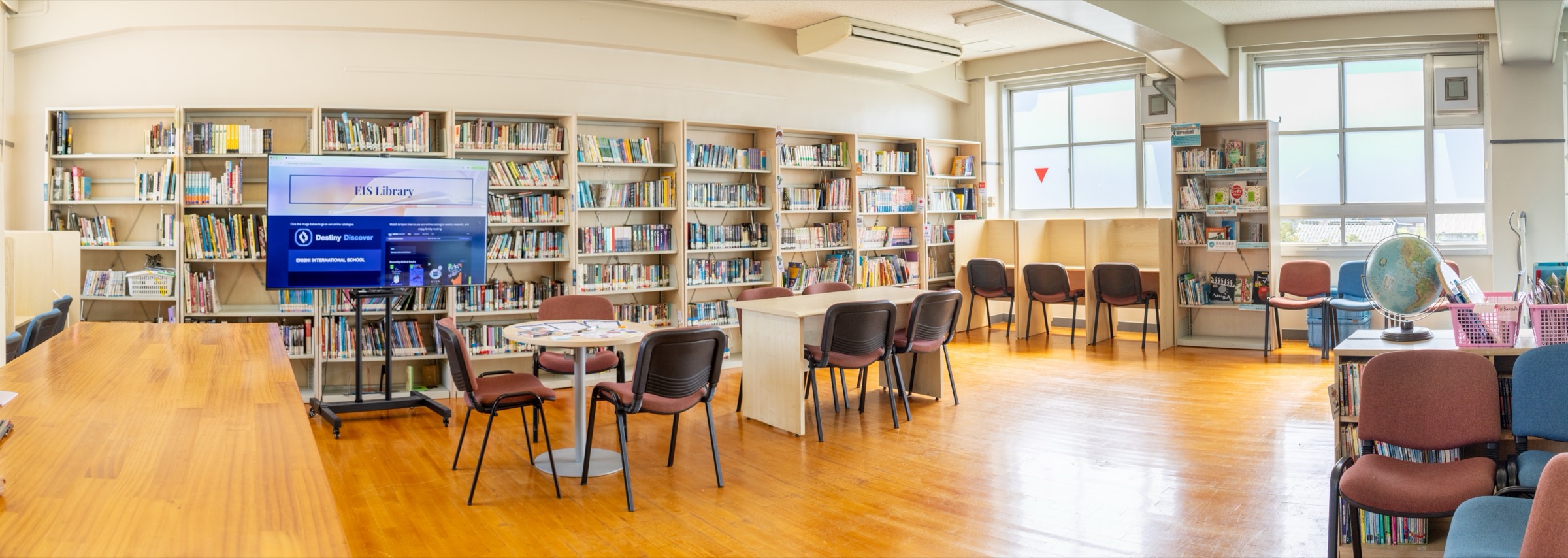 The Enishi library reading room: long wooden tables, low shelves of picture books and novels along the wall, a screen showing Destiny Discover, and a study corner by the windows.